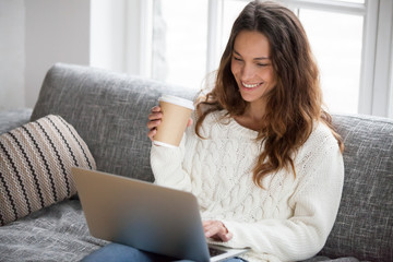Smiling young woman enjoying morning coffee using laptop sitting at home on sofa, attractive happy girl  typing on computer having fun online or chatting with friends while relaxing on couch
