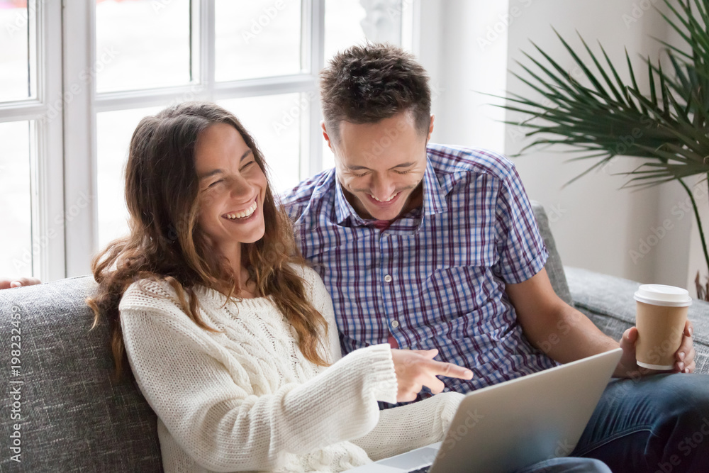 Wall mural Young happy couple watching funny video online, cheerful family having fun with laptop sitting on sofa at home, boyfriend and girlfriend friends laughing out loud at internet hilarious joke together