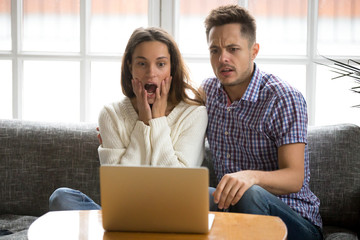 Shocked couple feeling confused and scared while watching horror scary film video movie online, worried young family sitting on sofa together looking at laptop screen with surprise or bewilderment