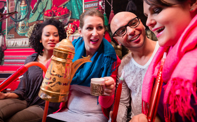 Group of friends smoking hookah and drinking coffee in shisha lounge enjoying themselves