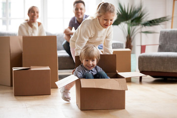 Happy siblings having fun moving into new home, smiling sister helping excited little boy riding in...