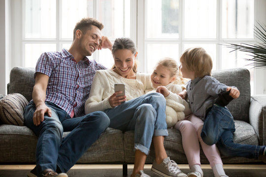 Cheerful young family with kids laughing watching funny video on smartphone sitting on couch together, parents with children enjoying playing games or entertaining using mobile apps on phone at home