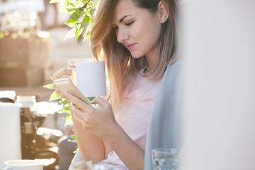 Woman enjoys morning coffee, using smart phone and relax. Beautiful sunny day, backlight