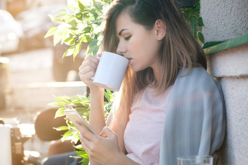 Woman drinking coffee, using smart phone and enjoying beautiful sunny day. Chilly morning, backlight