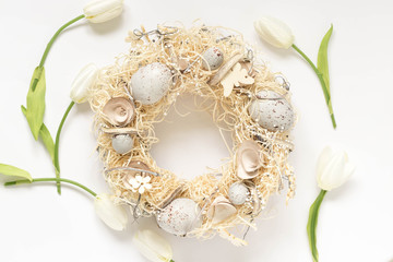 Easter wreath with straw, eggs and other traditional elements and white tulips around. Top view image of a pastel colored hand made home decoration for the spring holidays.