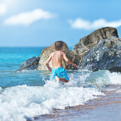 Cute Caucasian boy is running in the water along the sea shore. Back view.