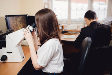 Student young woman sits behind a microscope in classroom, in background male student. Concept team work in scientific project.