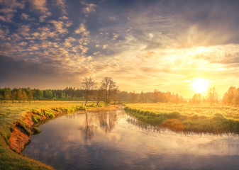 Nature landscape of spring river on morning dawn. Bright sun lights on green grass and young foliage. Rural scene. Trees on shore of river and colorful sky with clouds shining on warm sunlight.