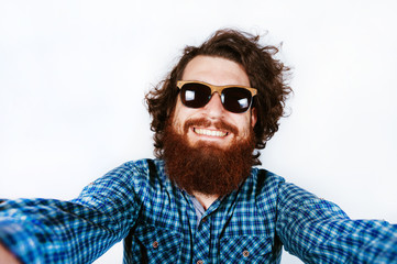 Obraz premium Cheerful happy bearded man with sunglasses looking at camera and taking selfie on white background in studio. Curly hair. Smiling young man with beard.