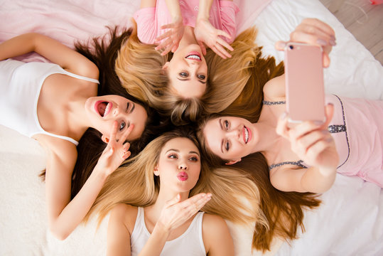 Sexy, pretty, charming, funny, foolish, lovely,  cheerful girls lying head to head on bed using smart phone for selfie, sending air kiss with pout lips, palm, gesturing peace symbol, two fingers