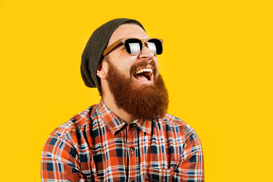 Portrait of young hipster man in sunglasses and hat posing on yellow background. Smiling bearded man wearing sunglasses, studio shot. Happy man with beard looking up.