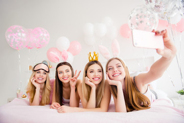 Cheerful, joyful, charming, pretty, funny, dreamy, cute models lying on bed shooting self portrait on front camera, holding using smart phone, showing two fingers, peace symbol, enjoying theme party