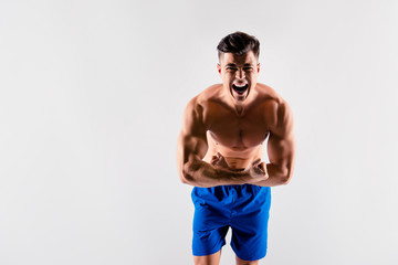 Portrait of aggressive screaming handsome athlete wearing blue loose shorts, he is demonstrating and toughening his muscular body, isolated on grey background