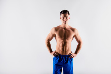 Fototapeta premium Portrait of sexy handsome muscular confident serious self-assured sportive athlete dressed in blue sportwear, he is staying still with hands on hips, isolated on grey background