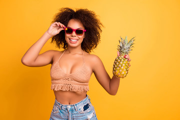 Carefree fooling around cheerful delightful wearing casual summer outfit and denim shorts african beautiful woman with curly brunette hair is going to eat pineapple, isolated on yellow background