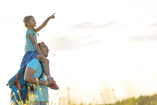 What Is It. Curious Little Girl Is Pointing Finger To Sky While Sitting On Her Father Shoulder. Man Is Standing On Meadow While Carrying Backpack. Copy Space