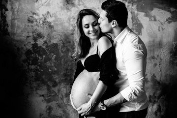 Happy couple. Pregnant woman. Black and white photo on which husband hugs and kisses his beloved pregnant wife in a black lingerie near a wall