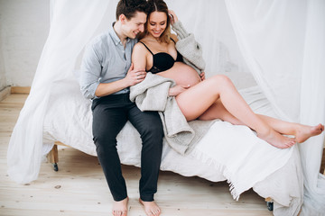 Happy cheerful couple. The husband hugs his pregnant beautiful wife, they are sitting together on the white bed in the bedroom