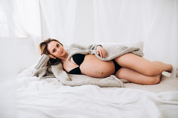 Naklejka premium Pregnant woman. Beautiful blonde pregnant woman in black lingerie with charming smile lies on the bed in the bedroom