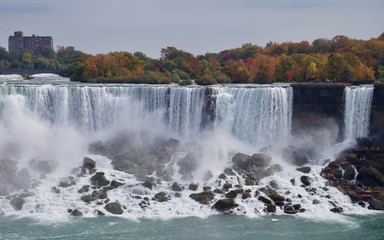 Fototapeta premium Beautiful and impressive panorama of the Niagara Falls in Ontario (Canada) on a bright autumn day with water crashing down the falls onto rocks creating lots of mist in front of the city skyline