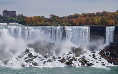 Beautiful and impressive panorama of the Niagara Falls in Ontario (Canada) on a bright autumn day with water crashing down the falls onto rocks creating lots of mist in front of the city skyline