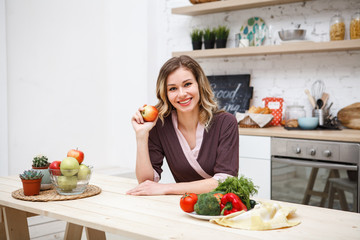 Beautiful blond girl in dressing gown sits at a kitchen table in a dining room in a hand holds an apple looking at the camera and smiling. Space for text. Vegetarianism.