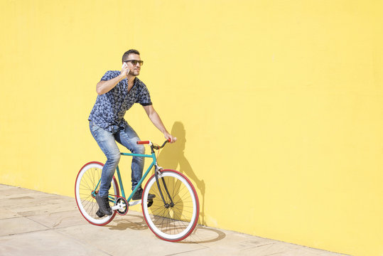 Man talking on smartphone while riding bicycle against yellow wall