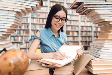 Ethnic asian girl sitting at table surrounded by books in library. Student is writing in notebook.