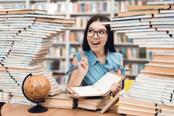 Ethnic asian girl sitting at table surrounded by books in library. Student is writing in notebook.