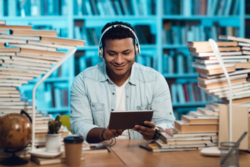 Ethnic indian mixed race guy surrounded by books in library. Student is using tablet.