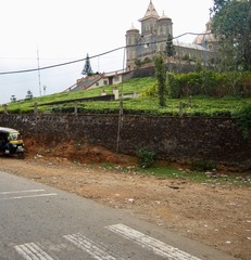 Rural Kerala (South India) with a tuk tuk / motor rickshaw parking on a dirt path next to a street in front of a green hill with an ancient temple on a cloudy day with grey sky