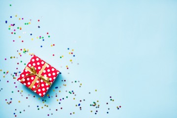 Obraz premium Top view of a red dotted gift box and scattered glittering star shaped confetti over blue background. Celebration concept. Copy space.