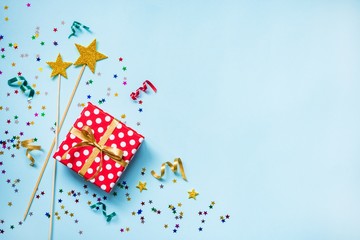 Top view of a red dotted gift box, golden magic wands, colorful confetti and ribbons over blue background. Celebration concept. Copy space.