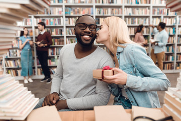 Ethnic african american guy and white girl surrounded by books in library. Students are giving gift.