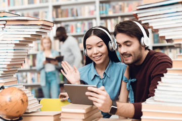 Ethnic asian girl and white guy surrounded by books in library. Students are using tablet with headphones.