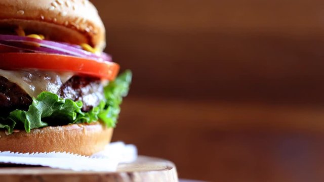 Grilled real American beef burger with lettuce, tomato, onion and mustard served on pieces of brown paper rotating on a rustic wooden counter. Close-up of burger with copyspace on wooden texture