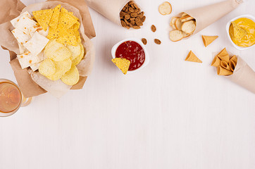 Crispy golden snacks on craft paper, triangles nachos, lager beer, red and yellow sauce in bowl on soft white wood background, with copy space, border. Summer fresh fast food background.