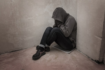Photo of desperate young drug addict wearing hood and sitting alone in corner - obrazy, fototapety, plakaty