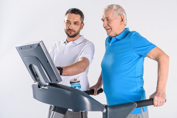 rehabilitation therapist assisting senior man exercising on treadmill isolated on grey