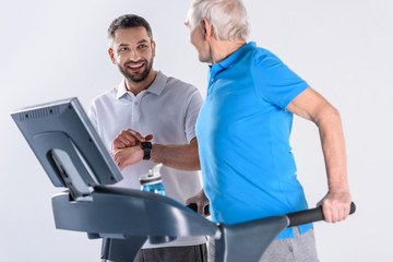 smiling rehabilitation therapist assisting senior man exercising on treadmill isolated on grey