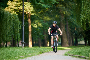 Toned male biker in professional sportswear and protective helmet looking to side while riding bike down park alley on summer day. Exercising, reaching goal
