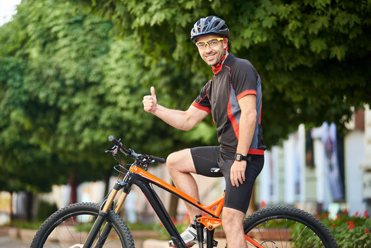 Happy professional bicyclist in cycling sportswear and protective helmet showing thumbs up sitting on bike in front of green trees and smiling to camera. Sportsman satisfied after morning training