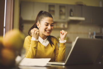 Happy young woman sitting at home and working on laptop and receiving good news.