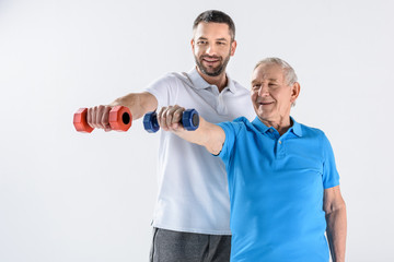 portrait of smiling rehabilitation therapist and senior man exercising with dumbbells isolated on grey