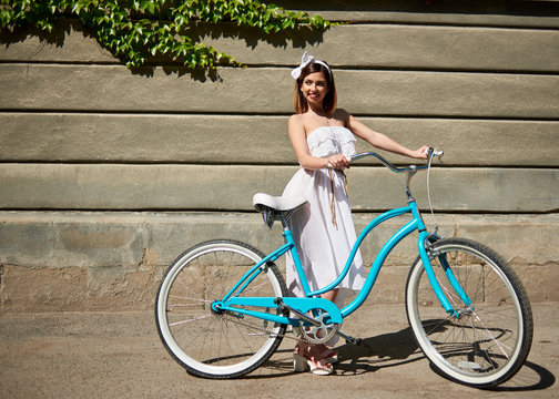 Smiling girl in white summer dress with a blue vintage bicycle looking away on a wall background with green creeper at sunny day