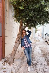 Photo of a smiling Caucasian girl with long hair in full length. A girl with long hair in a denim suit and white sneakers leaned her elbows on the olive tree. Summertime