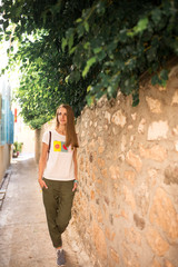 Caucasian girl in white T-shirt with fruits, green pants and gray sneakers is walking along a narrow ancient street with trees and blue shutters in the windows of the houses.