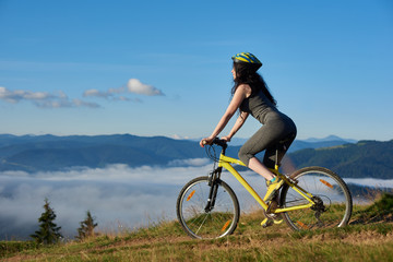 Obraz premium Back view of sporty girl cycling on yellow bike on rural trail in the mountains, wearing helmet, on sunny morning. Foggy mountains, forests on blurred background. Outdoor sport activity. Copy space