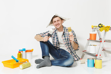 Smiling young man in newspaper hat with brush, putty knife instruments for renovation apartment room isolated on white background. Wallpaper, gluing accessories, painting tools. Repair home concept.