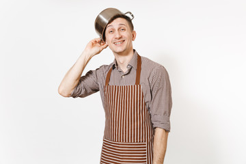 Fun man in striped apron with silver stainless glossy aluminium empty stewpan, pan or pot on head isolated on white background. Male housekeeper or houseworker. Kitchenware, dishes, cuisine concept.